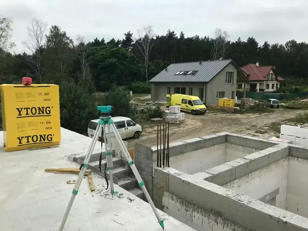 Niwelator geodezyjny na statywie na płycie fundamentowej, materiały YTONG i van na placu budowy, etap pomiarów