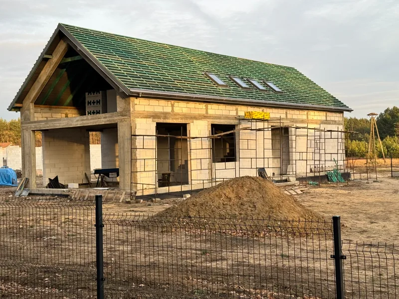 Parterowy dom jednorodzinny ze ścianami z gazobetonu H+H z dachem pokrytym kontrolątami i zieloną folią wysokoparoprzepuszczalną, zamontowane okna dachowe Velux, rusztowania na elewacji, złota godzina, leśna lokalizacja