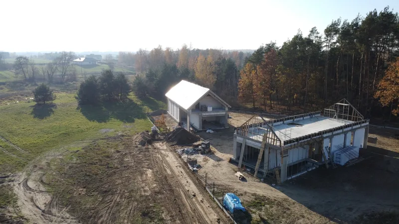Lotnicze zdjęcie dwóch domów w różnych fazach budowy — parterowy w stanie surowym zamkniętym z białym dachem dwuspadowym oraz drugi w fazie wieńca żelbetowego z otwartą więźbą, jesień, leśne otoczenie, aglomeracja warszawska